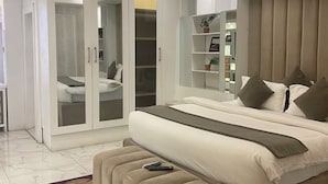 A modern bedroom with a bed, a wardrobe with glass doors, a bedside table, and a large mirror. - The Palms Hotel (Abuja)
