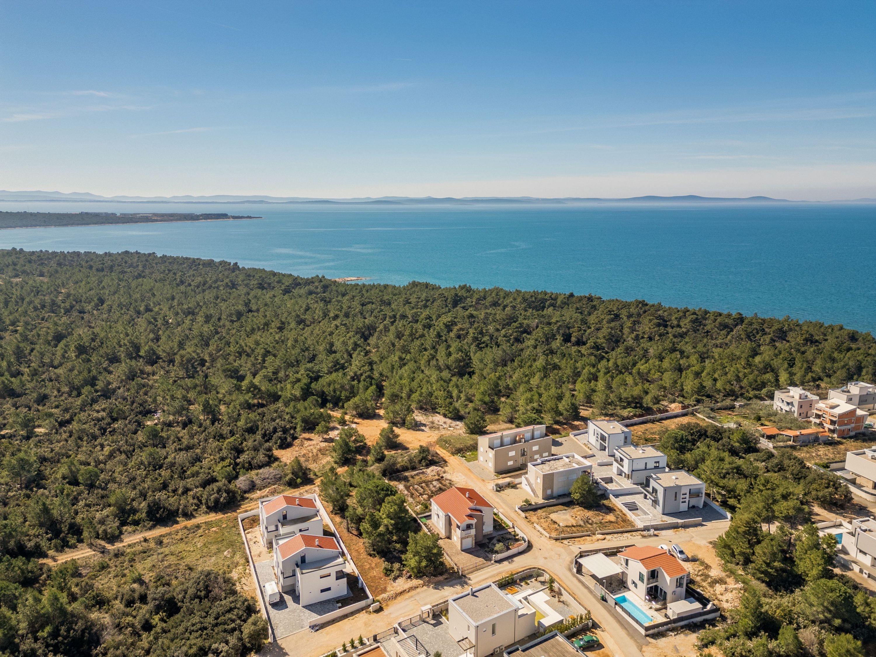 An aerial view of a coastal residential area with modern houses, a clear blue sea, and a dense forested hillside.