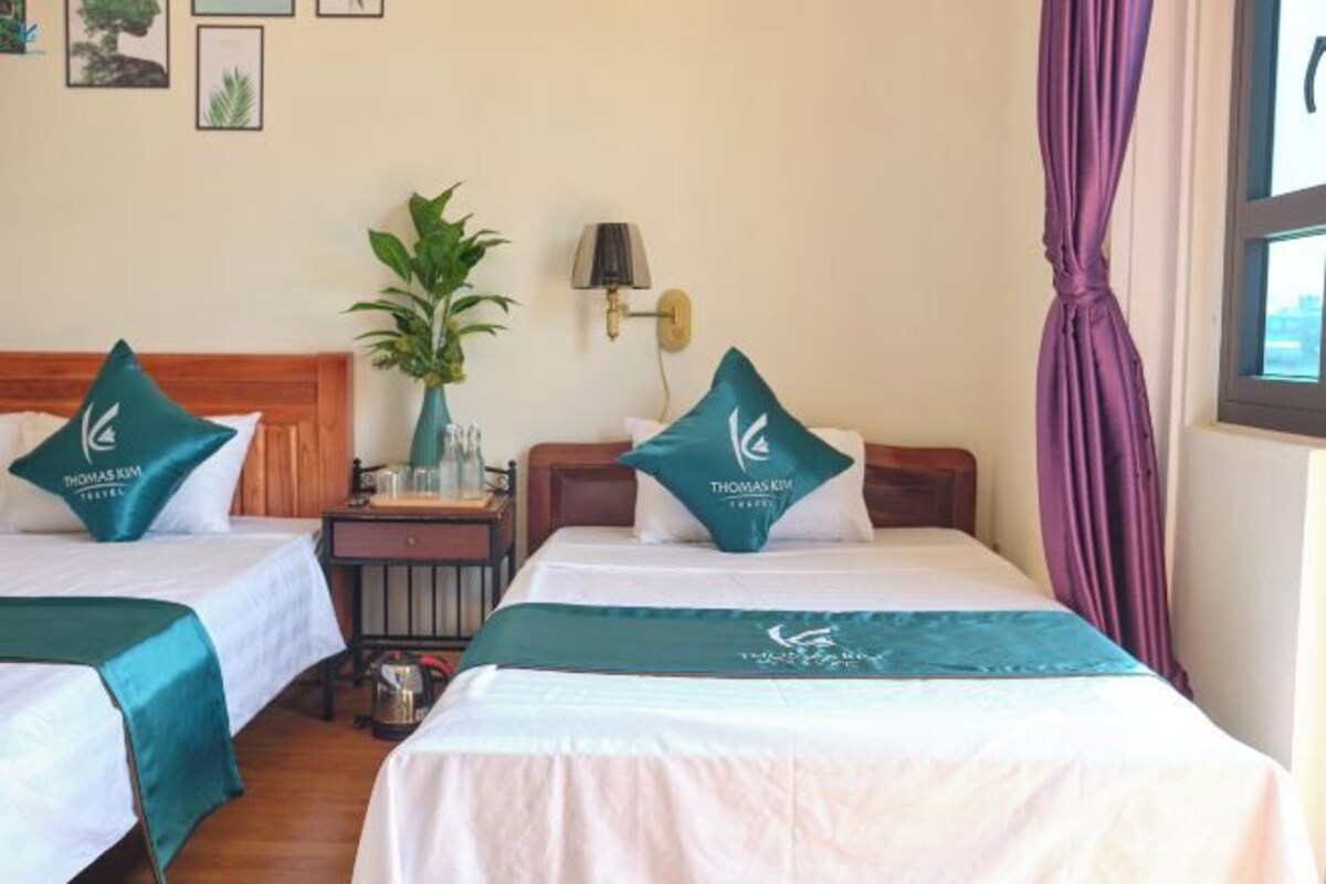 A hotel room with two beds, each with a teal bedspread and matching pillows, a nightstand with a lamp, a potted plant, and framed pictures on the wall.
