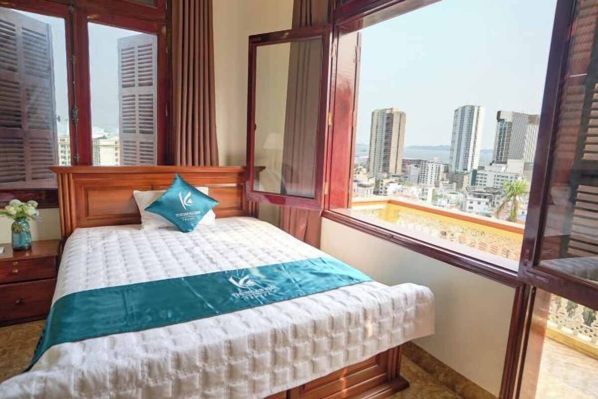 A hotel room with a bed, a window with a view of tall buildings, and a wooden headboard.