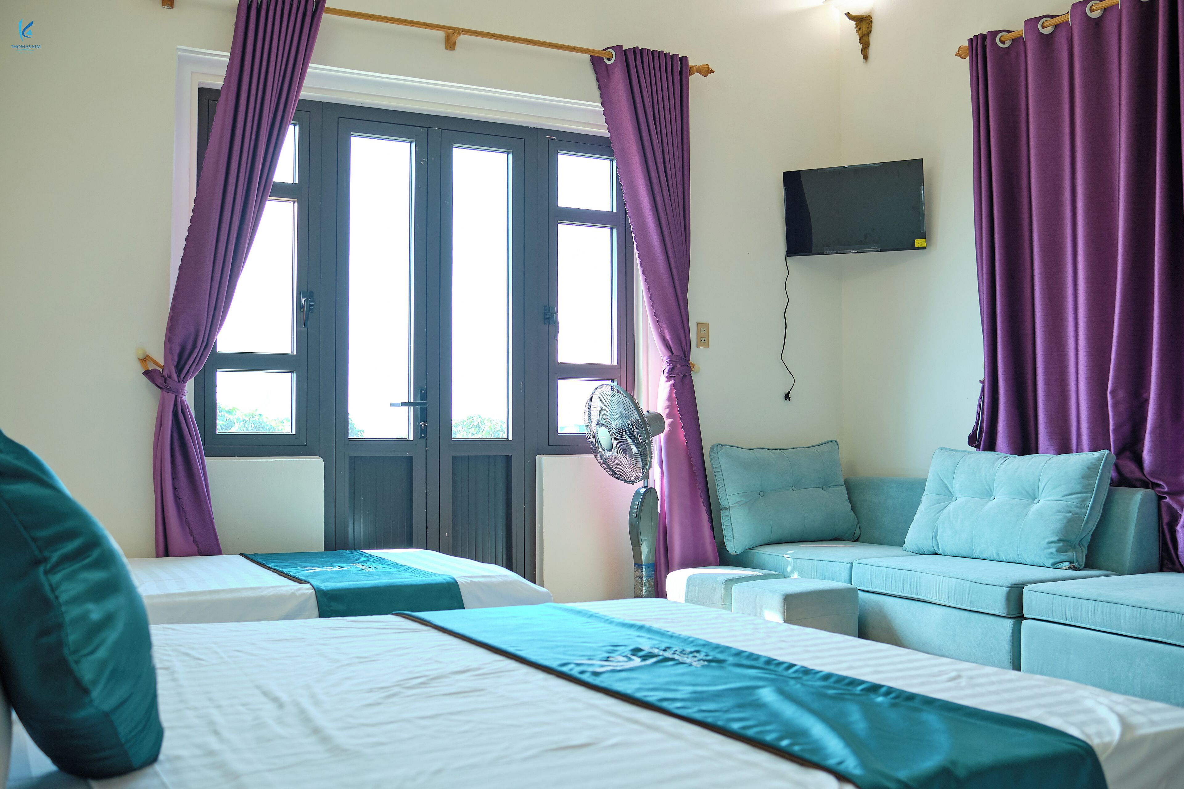 A hotel room with two beds, a sofa, a TV, and a window with purple curtains.