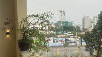 A balcony with a potted plant, a wall sign reading "ALL DAY COFFEE", and a view of a cityscape with buildings and a street.