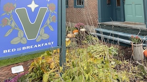 A sign for The Vesper Bed and Breakfast with a large 'V' logo, a brick building with blue trim, and a garden with various plants and pumpkins. - The Vesper  (Woodstock)