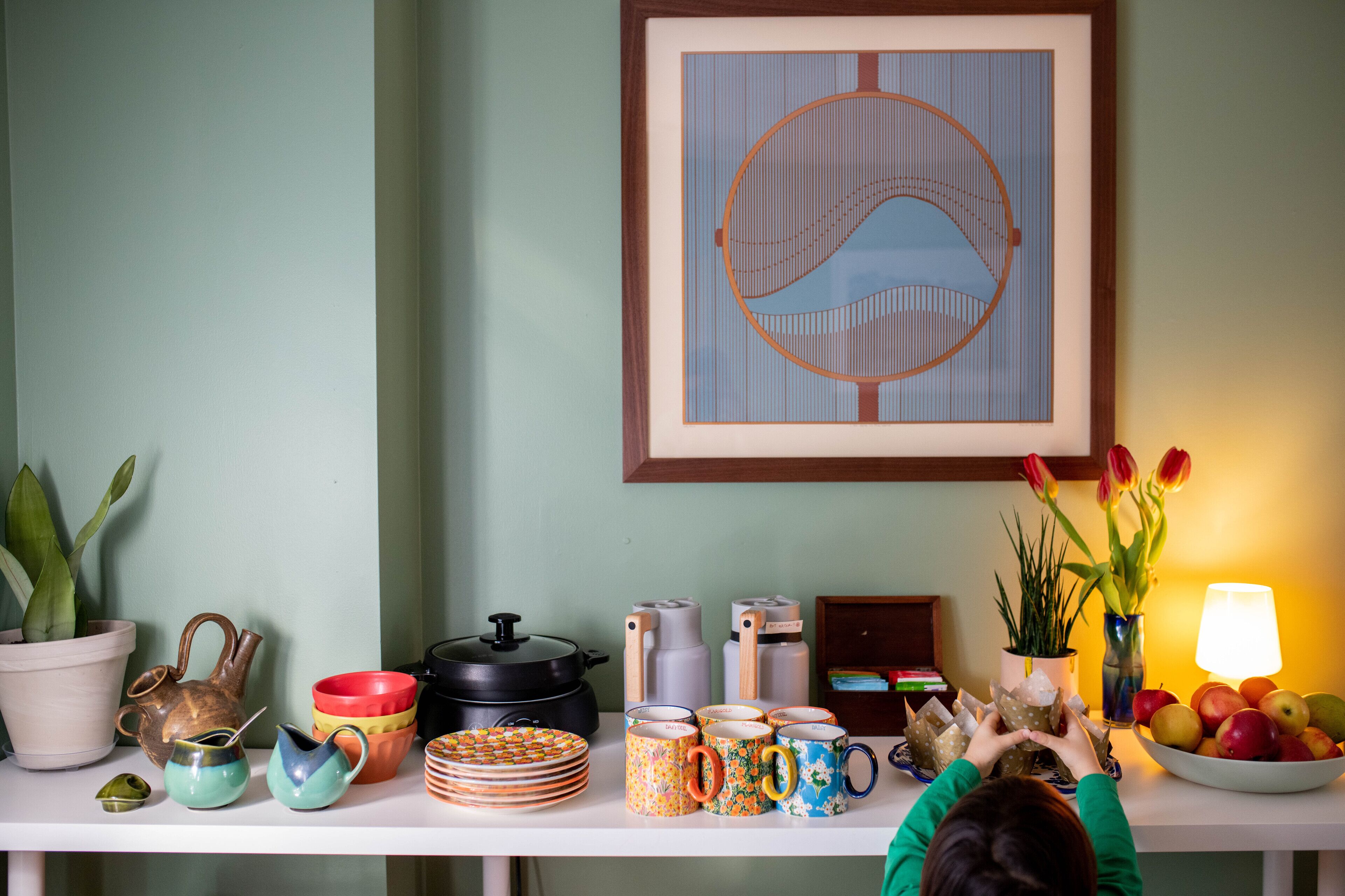 Uma mesa de jantar com utensílios de cozinha, uma planta em vaso e uma obra de arte emoldurada.