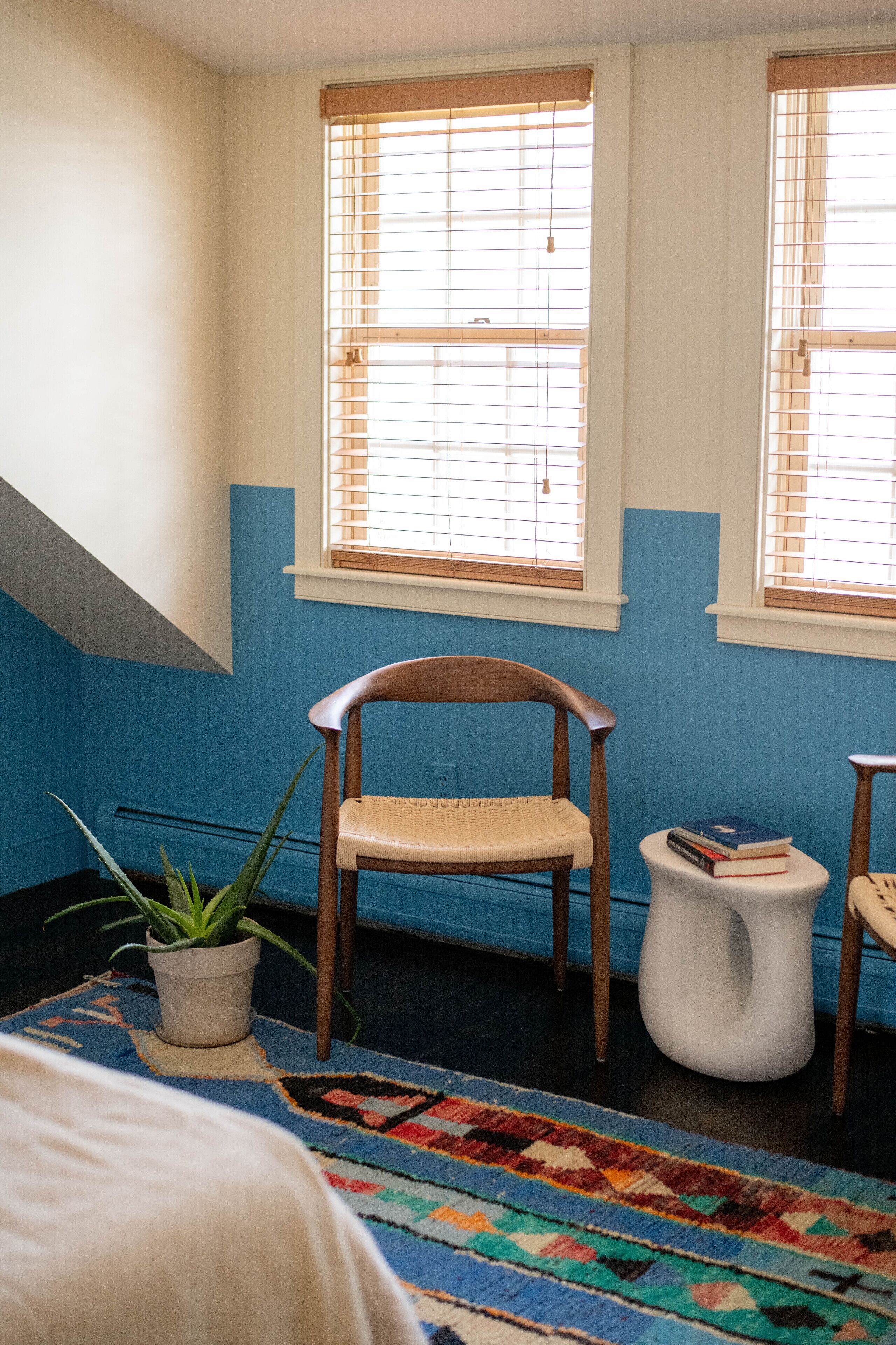 A room with a sloped ceiling, two windows with blinds, a wooden chair, a small white stool, a potted plant, and a colorful rug.