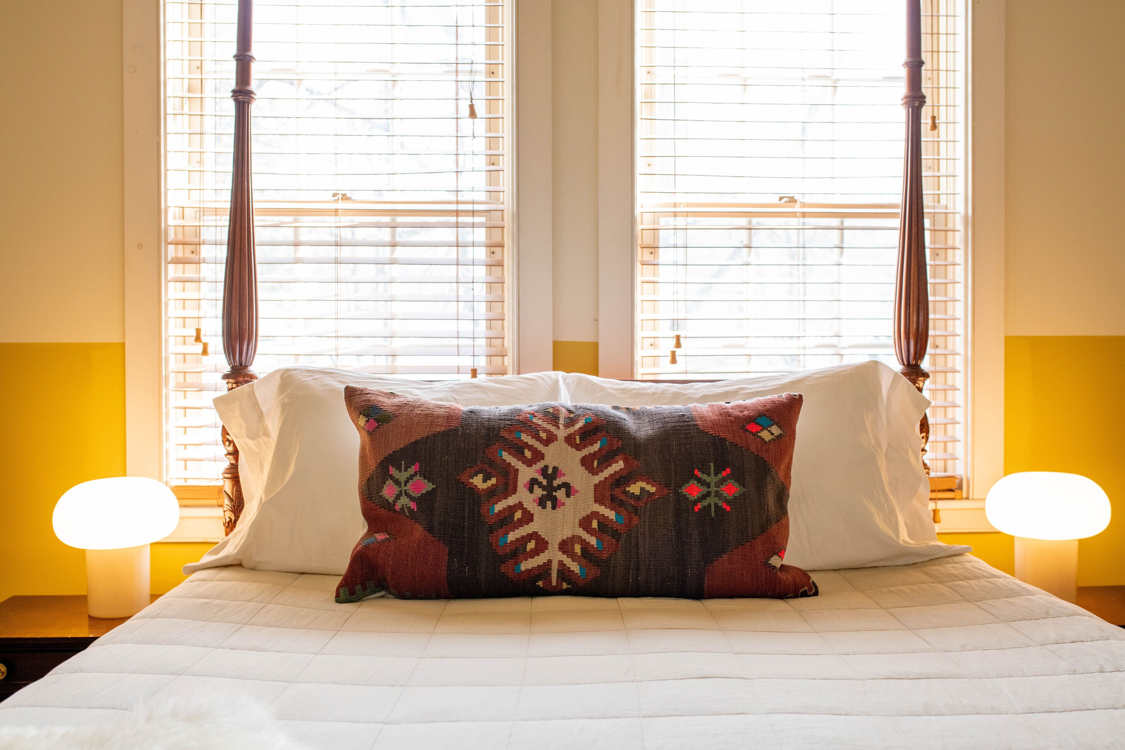 A bed with a patterned pillow, two bedside lamps, and a window with blinds.