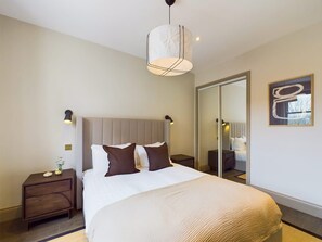A hotel bedroom with a large bed, a bedside table, a nightstand, a wall-mounted lamp, and a mirror. - Bright & Stylish Home by Abbey Road Studios (Greater London)