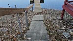 Un escalier en béton mène à une jetée en bois qui surplombe un lac paisible, entouré de feuilles mortes et bordé, sur la droite, d’une structure rouge.