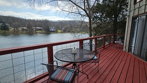 Une terrasse en bois avec une table et des chaises en métal, donnant sur un lac où l’on aperçoit des maisons au loin.