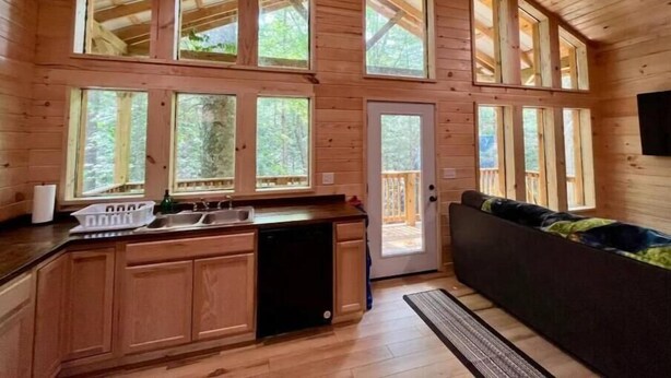 Una cabaña con zona de cocina que incluye armarios de madera, un lavabo y un lavavajillas. Varias ventanas ofrecen vistas del bosque circundante.