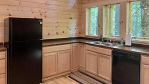 Una cocina con paredes de madera, un refrigerador negro, armarios de madera, lavavajillas, un fregadero con ventana arriba y una ventana con vista a árboles.