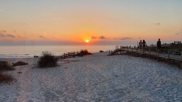 A beach with a wooden boardwalk leading to the water, featuring a sunset view.