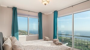 Une chambre avec un grand lit, deux fenêtres avec des rideaux et une vue sur la mer.