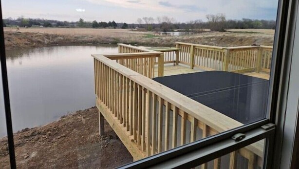Vista da interno che si affaccia su un pontile di legno con superficie nera, con vista su un'acqua calma e un paesaggio lontano.