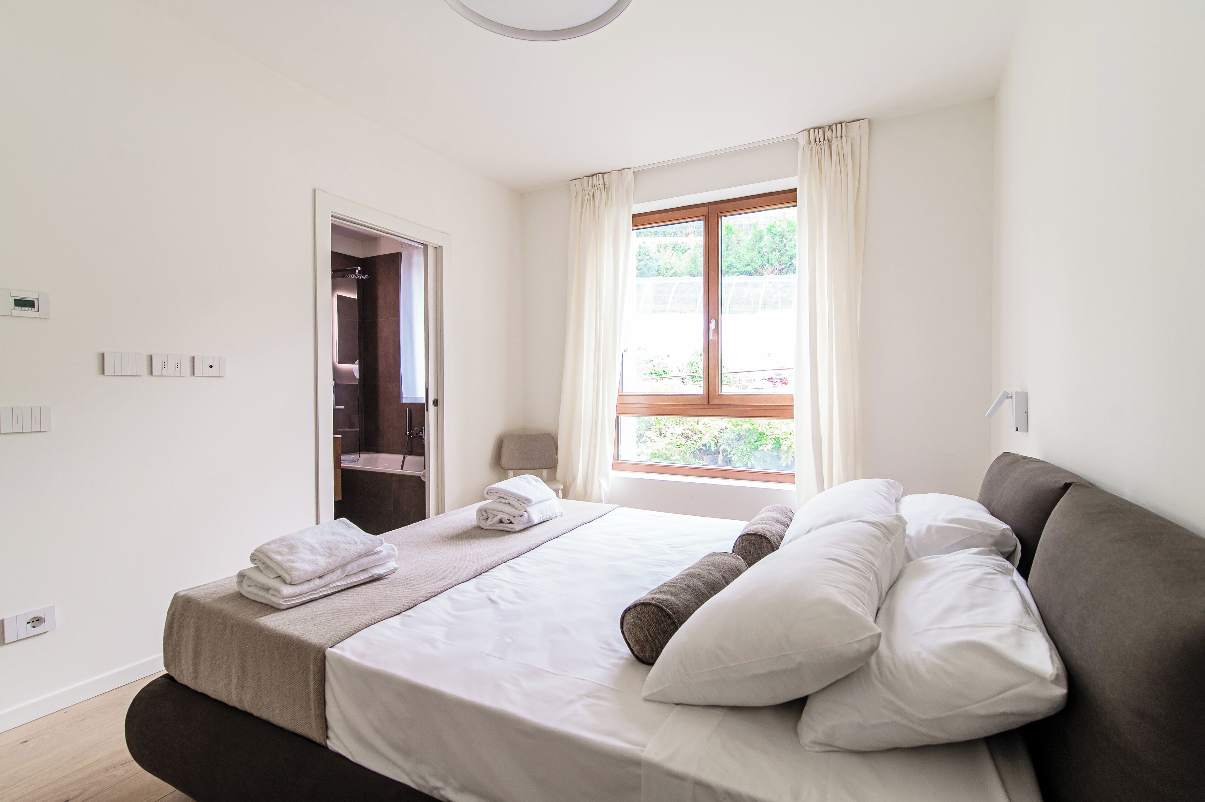 Una cama bien hecha con ropa blanca y un cabecero marrón, una ventana con cortinas y una puerta que da a un baño.