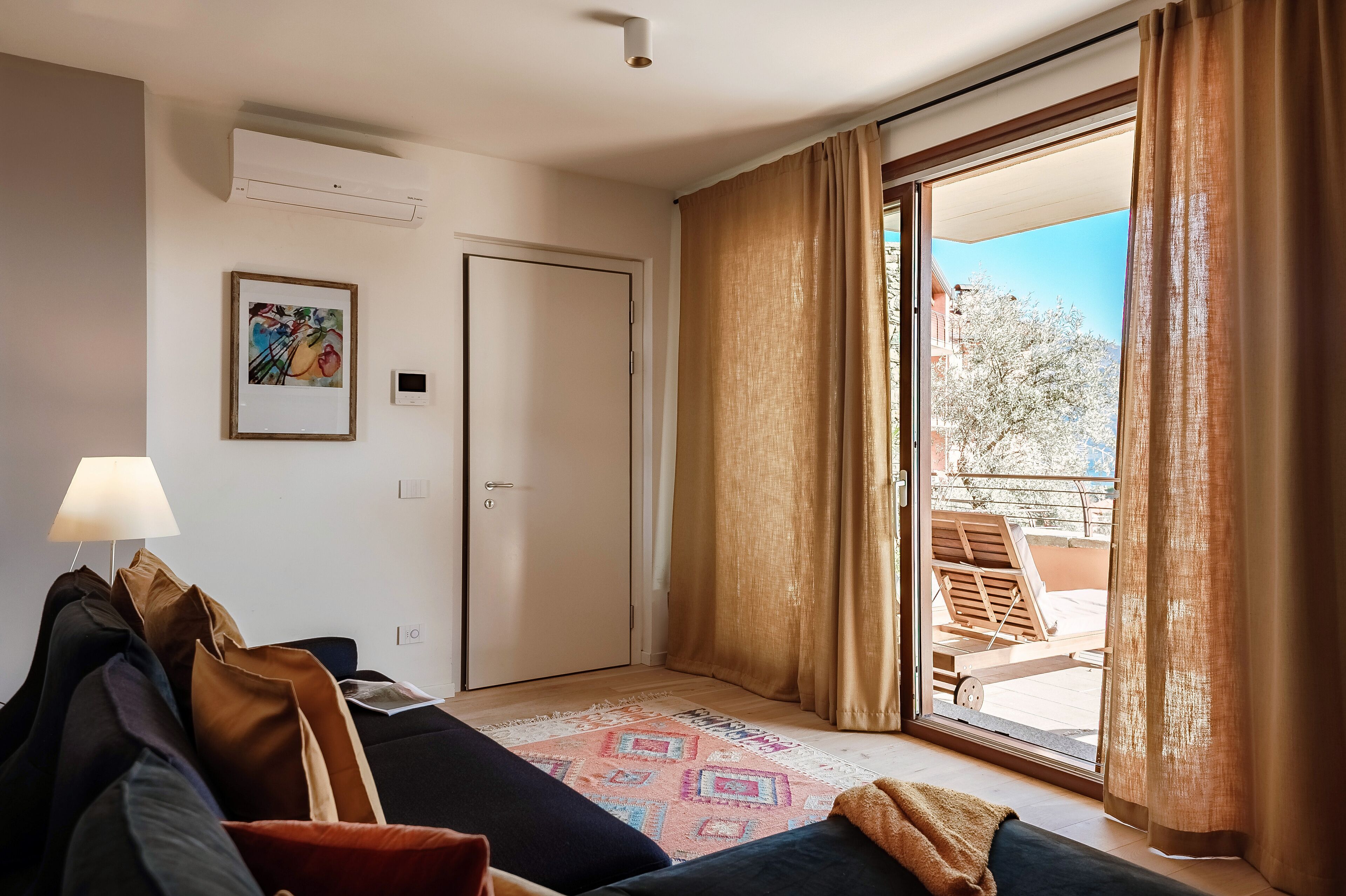 Une chambre d’hôtel comprenant un lit, un bureau, une lampe, un tableau et une porte-fenêtre coulissante donnant sur un espace extérieur avec une table et des chaises.