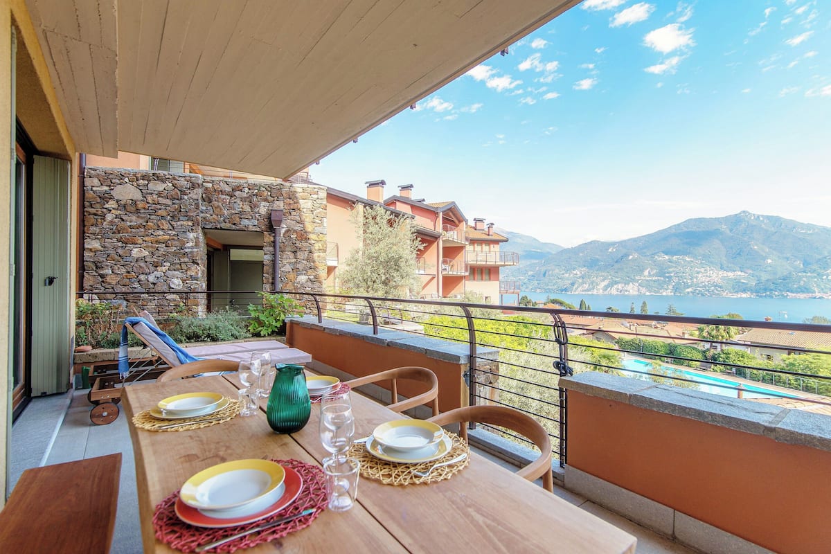 A balcony with a table set for dining, overlooking a mountainous landscape and a body of water.