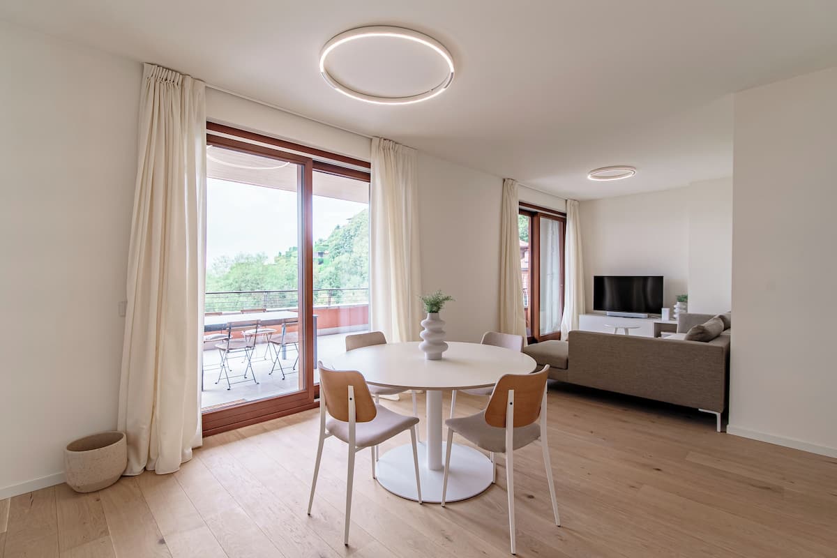A modern living room with a round dining table, two chairs, a sofa, a TV, and a balcony with outdoor furniture.