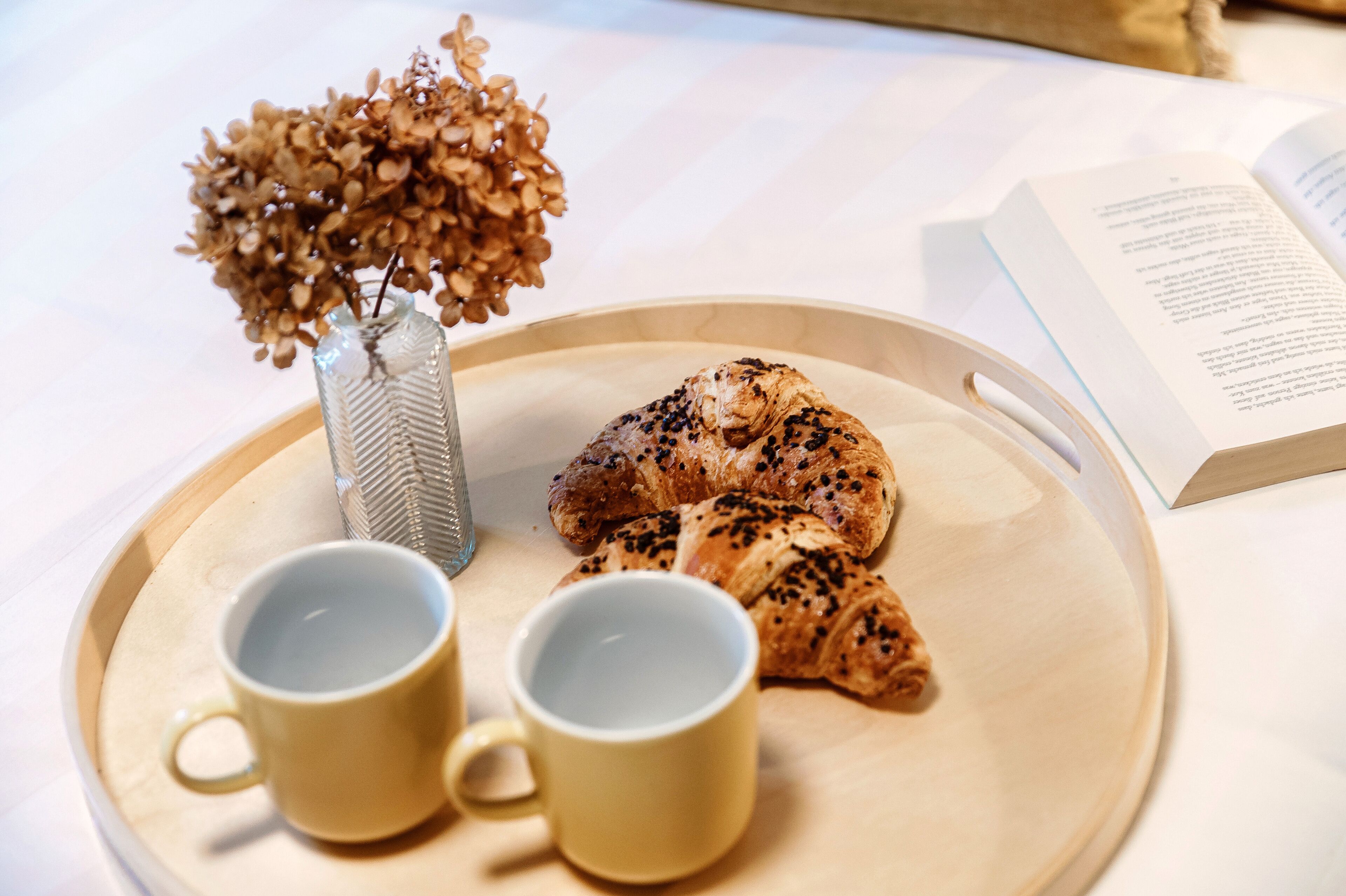 Una bandeja con dos tazas amarillas, un florero con flores secas y un libro sobre una mesa.