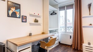Une chambre moderne avec un bureau en bois, une chaise, un radiateur, un chauffage d’appoint, une fenêtre donnant sur des immeubles et un rideau.
