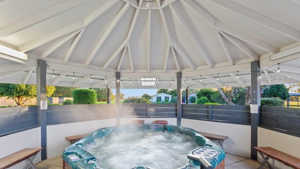 A hot tub under a white pavilion with benches, surrounded by greenery.