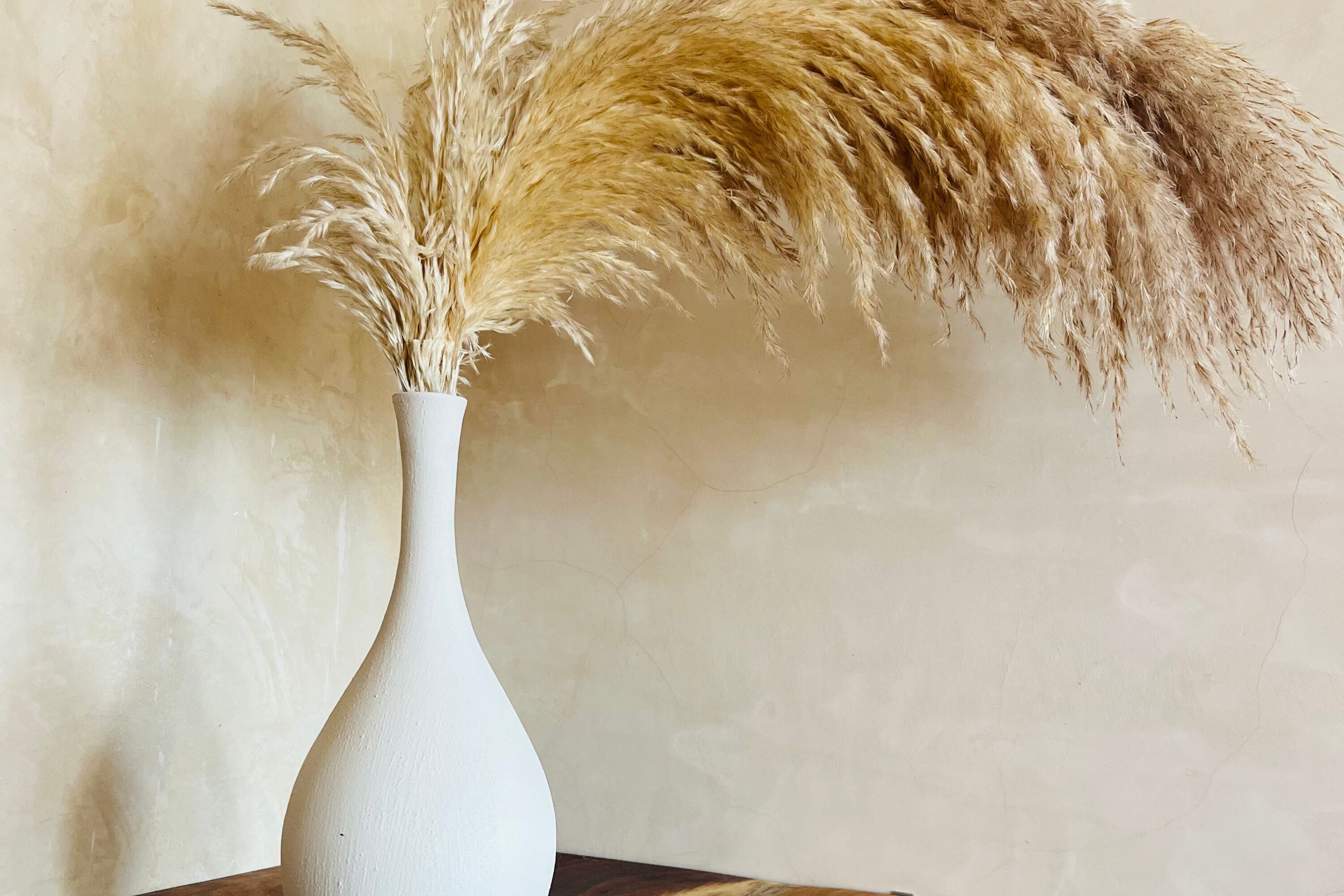Un vase blanc contenant des plumes d’herbe de la pampa, posé sur une table en bois devant un mur à texture.
