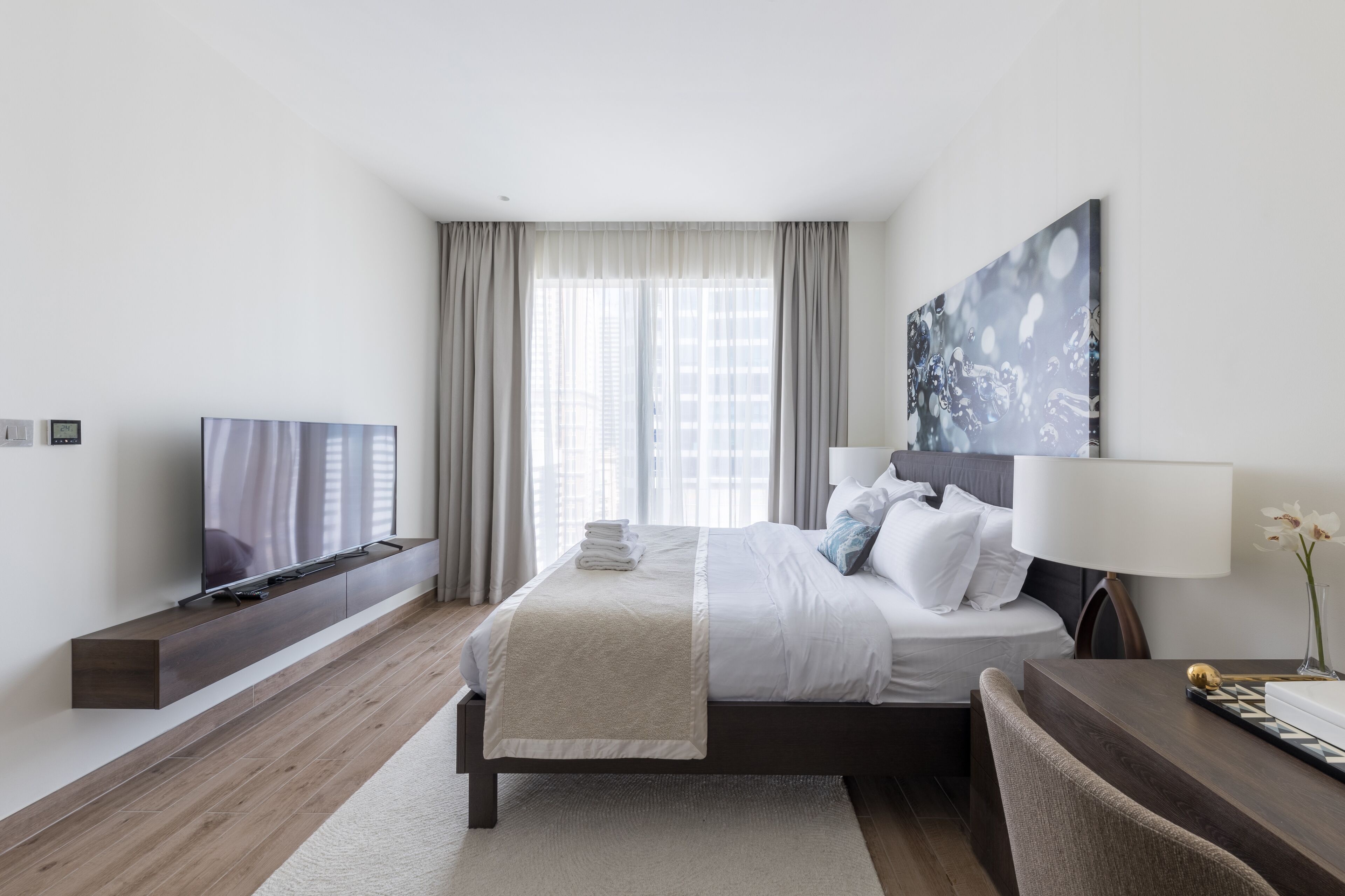 A modern hotel room with a bed, a TV, a desk, and a large window with curtains.