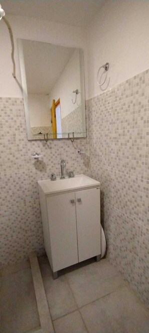 A bathroom with a tiled wall, a white sink with a mirror above it, and a small shelf. - casa nuestra hostal (Salta)