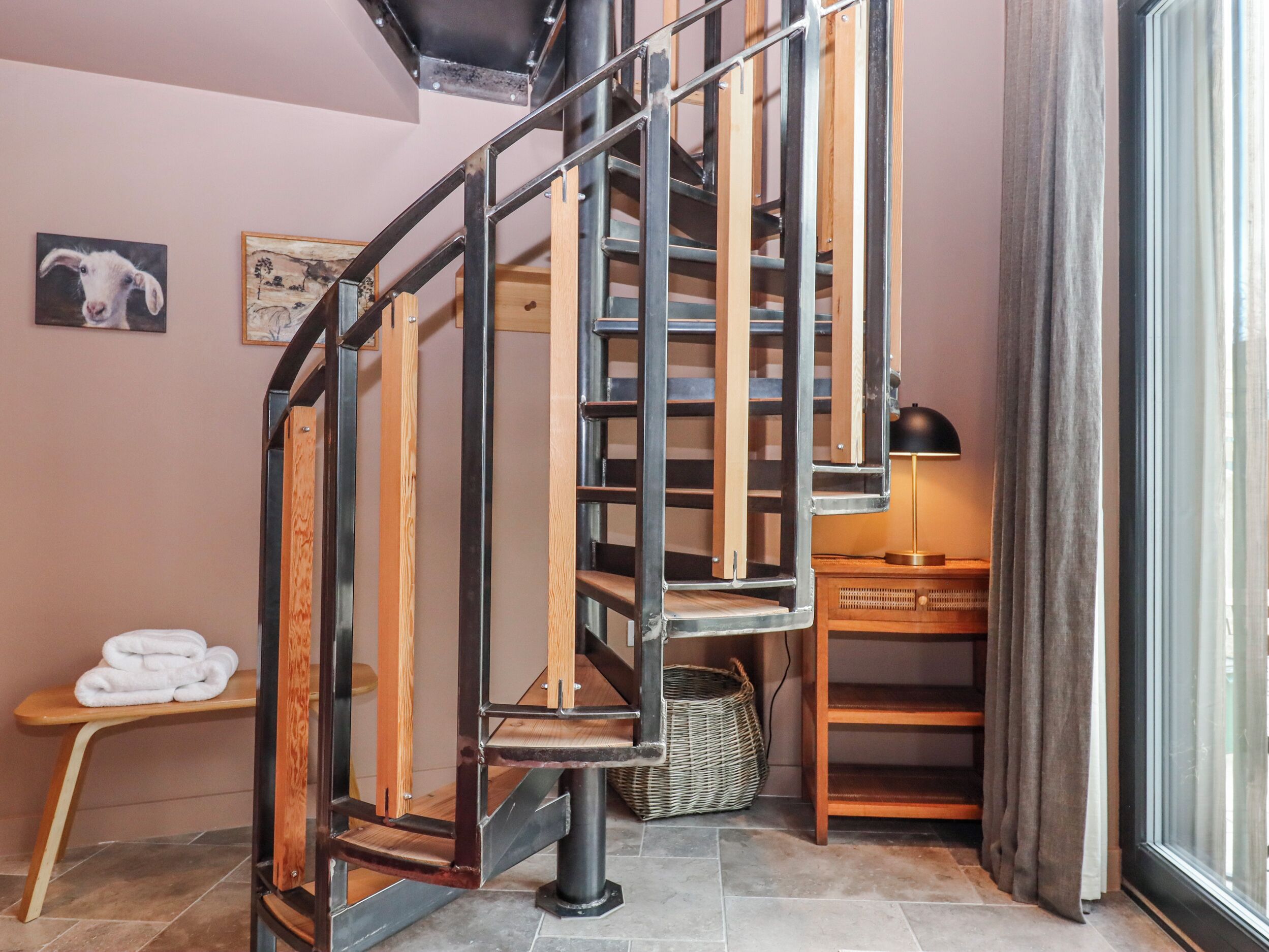 Una escalera de caracol con escalones de madera y barandillas de metal, una mesita con toallas y una cesta de mimbre.