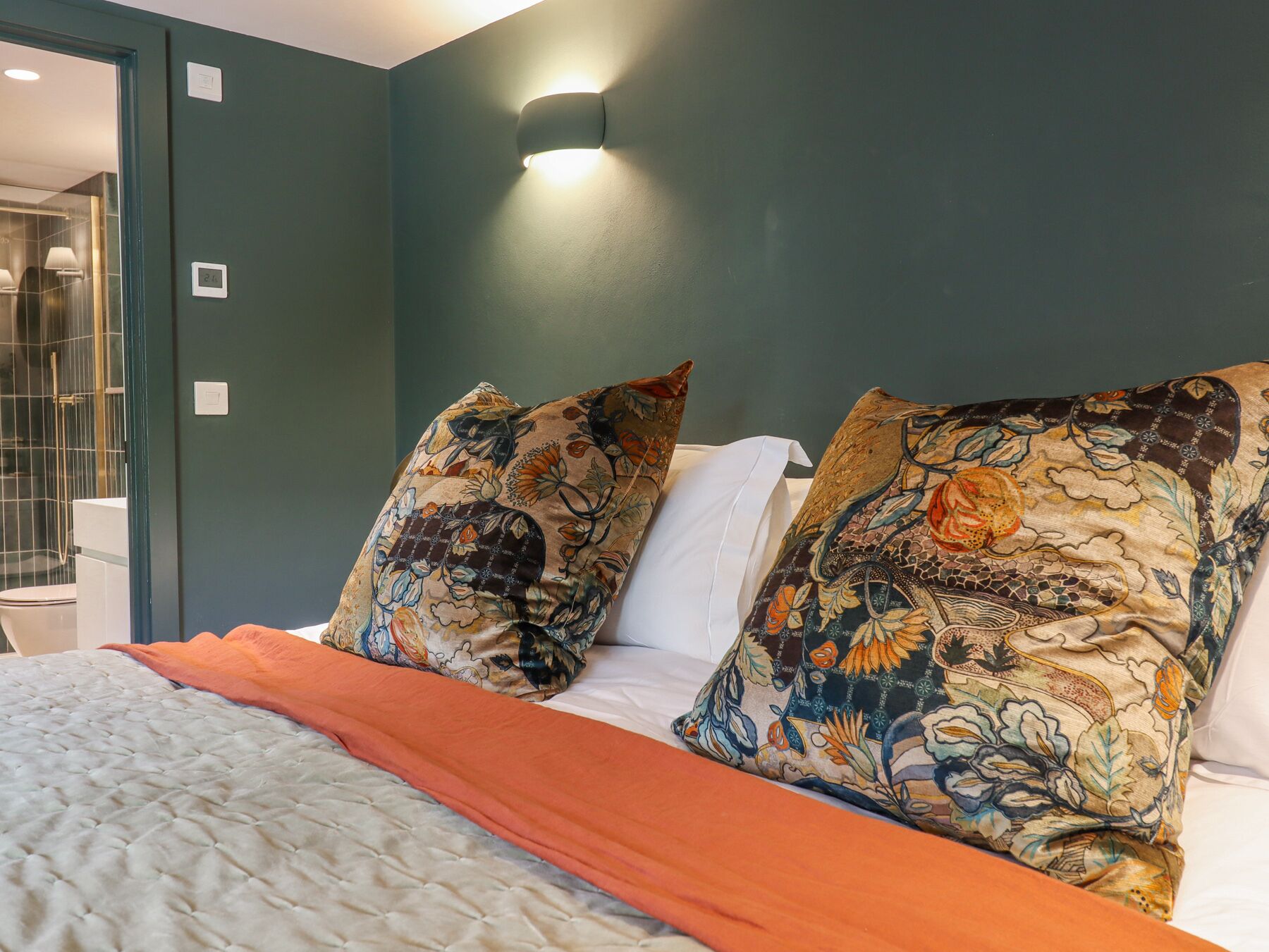 A bedroom with a bed, two patterned pillows, a white headboard, and a wall-mounted light fixture.