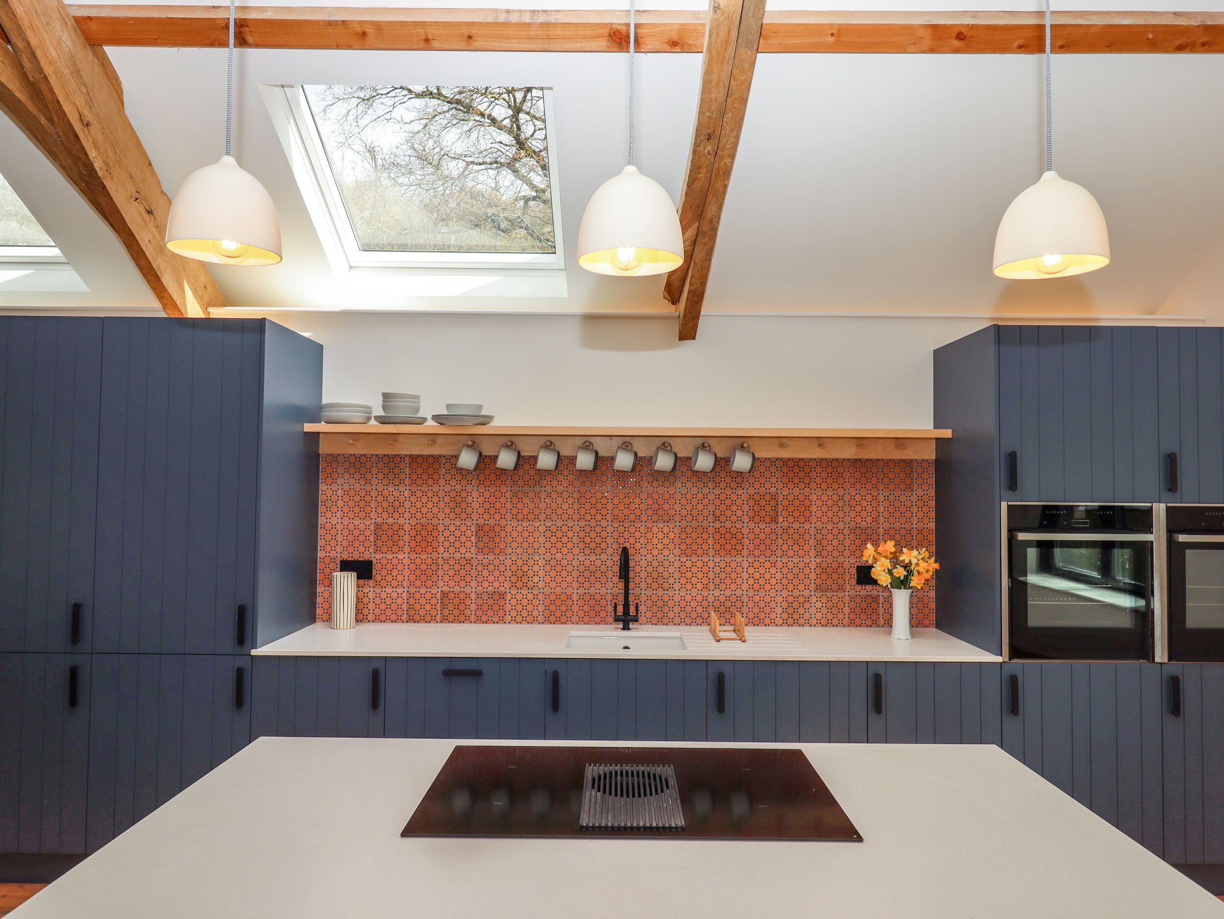 A modern kitchen with blue cabinets, a white countertop, a backsplash with orange and black tiles, and a skylight above.