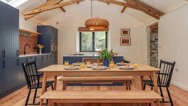 A modern kitchen with dark cabinets, a wooden dining table, and a stone accent wall. - 3 Caddaford Barns (Buckfastleigh)