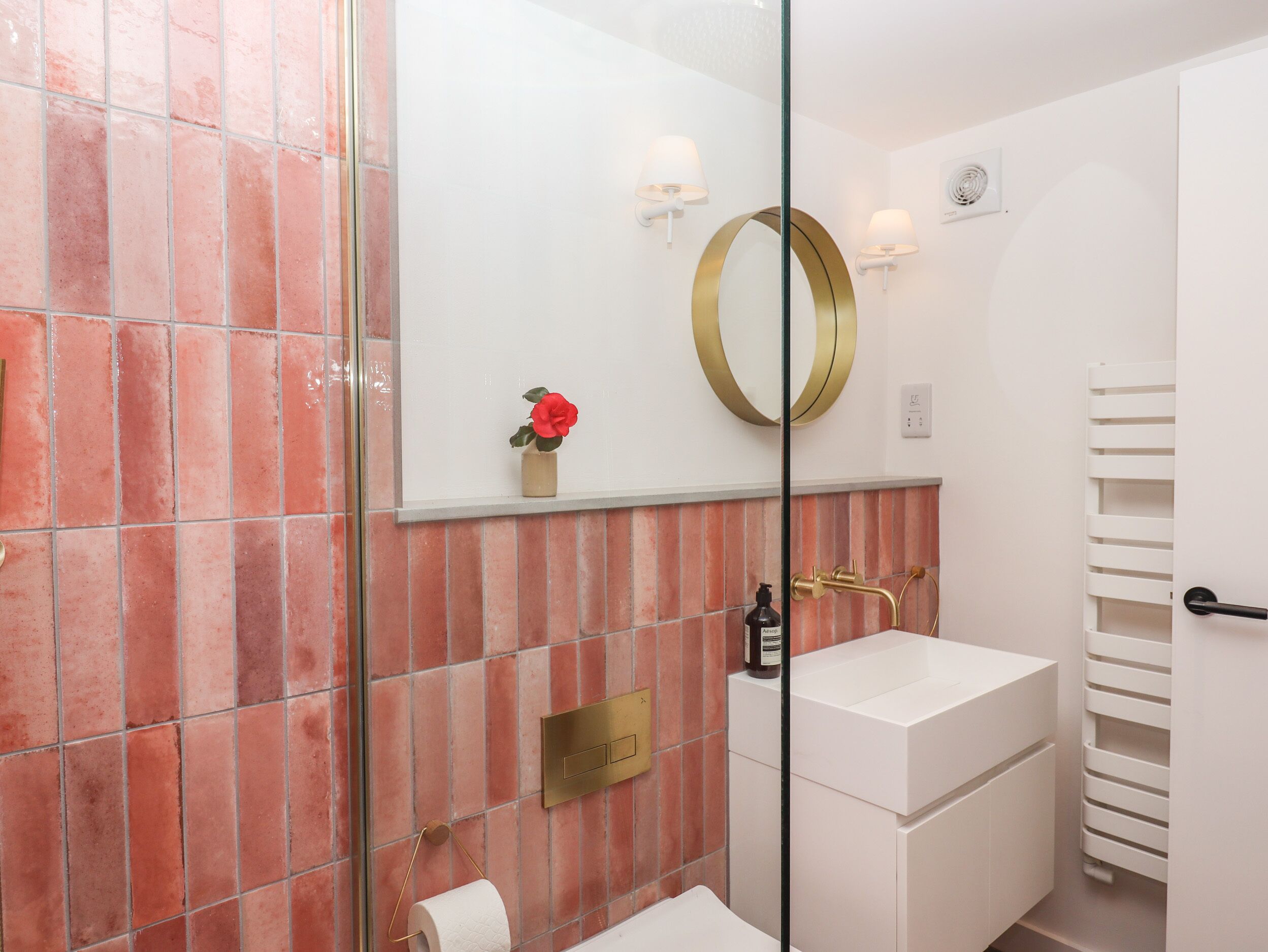 A modern bathroom with a white sink, a mirror with a gold frame, and a red flower vase.