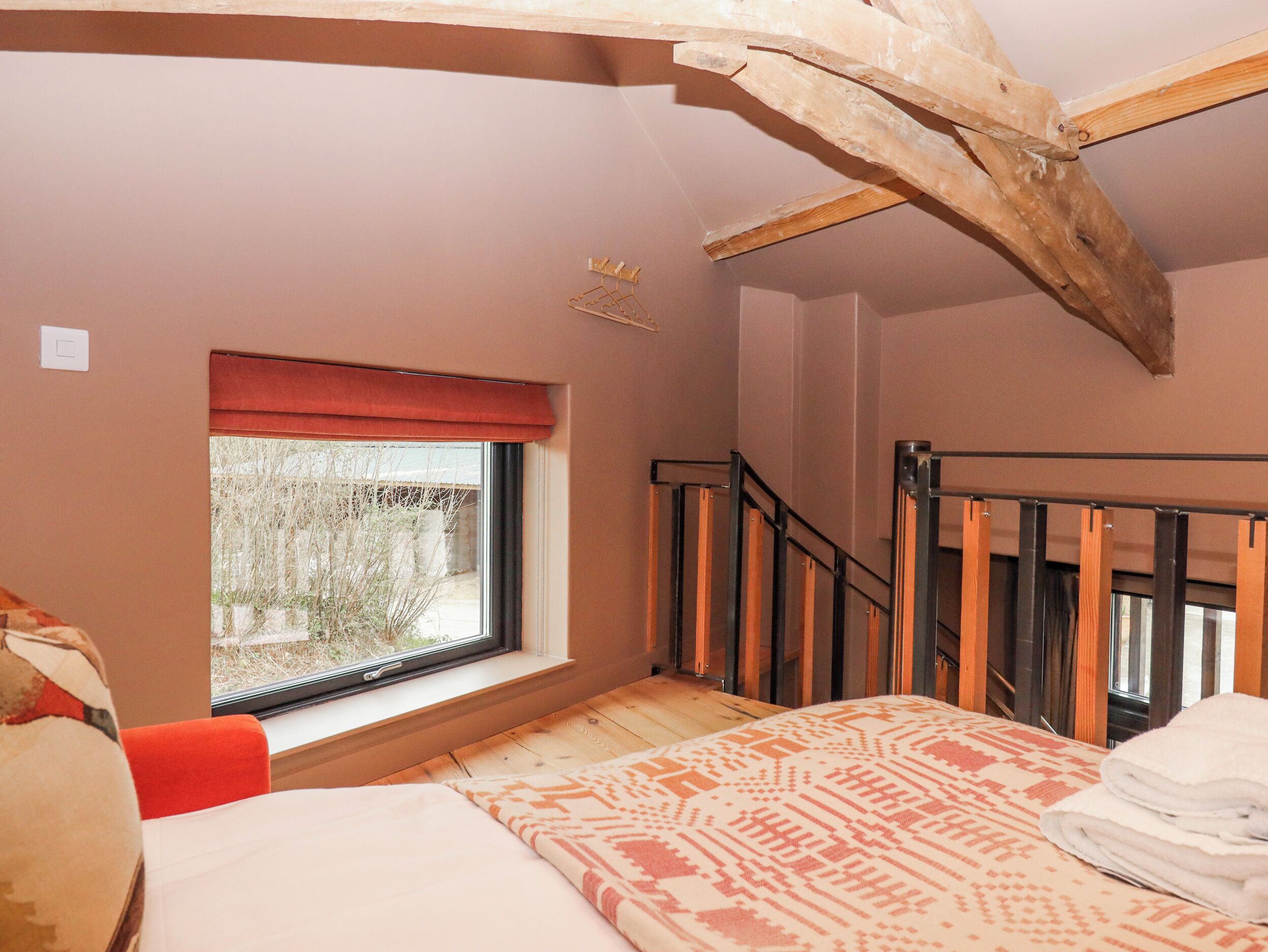 A bedroom with a window, a bed, a wooden ceiling beam, a staircase, and a view of trees outside.