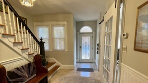 A spacious hallway with a patterned rug, a staircase with white balusters, and a chandelier. - Chester County Oasis with Pool, Modern Ammenities, Tranquil and Private (West Chester)