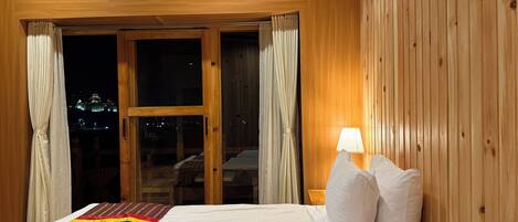 Habitación de hotel con interior de madera, cama con ropa de cama blanca, manta colorida y vista nocturna de la ciudad a través de un ventanal grande.