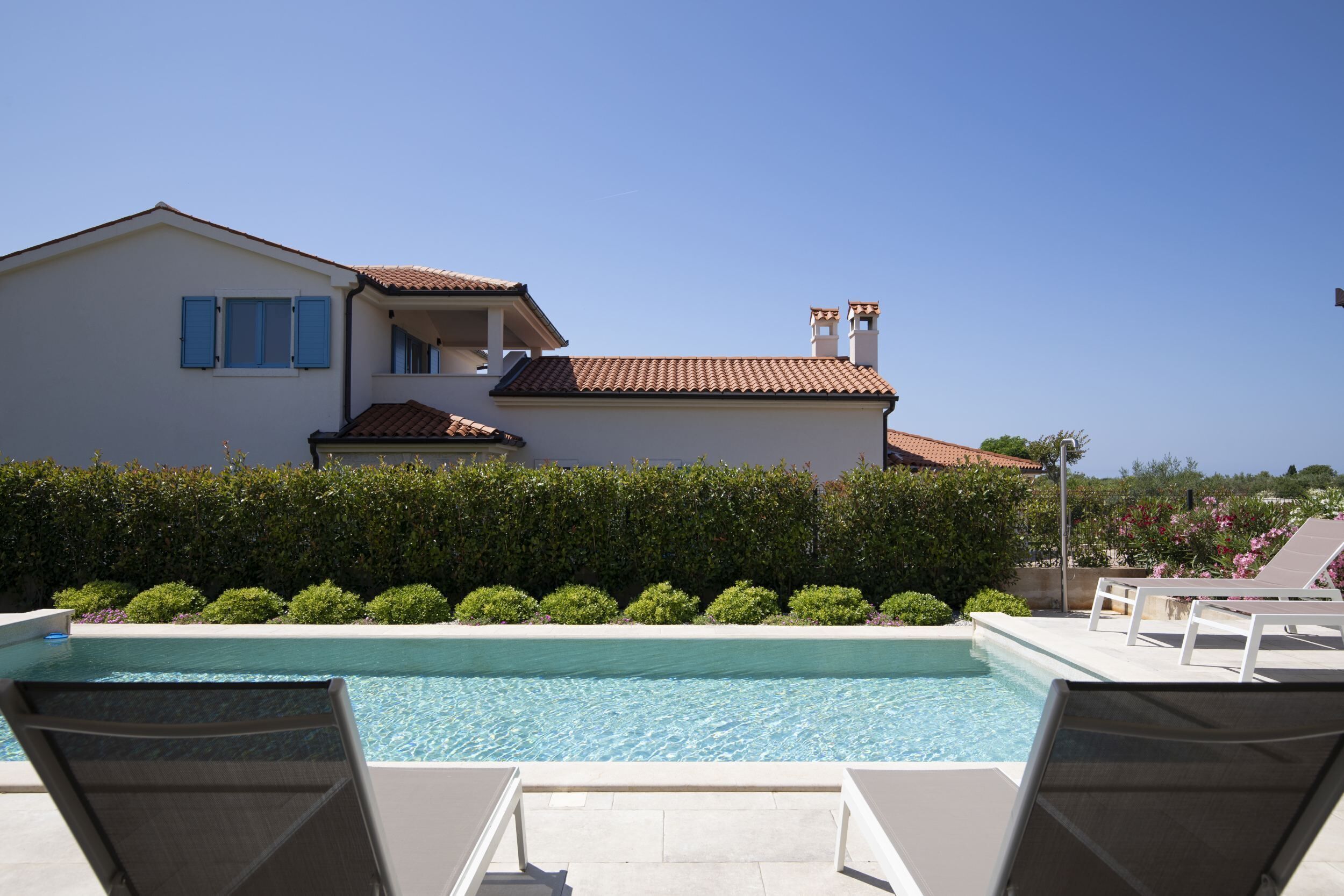 Une piscine avec des chaises longues, un bâtiment blanc aux volets bleus et un ciel d’un bleu limpide.