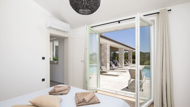 A bedroom with a bed, towels, and a sliding glass door leading to a pool area with lounge chairs.