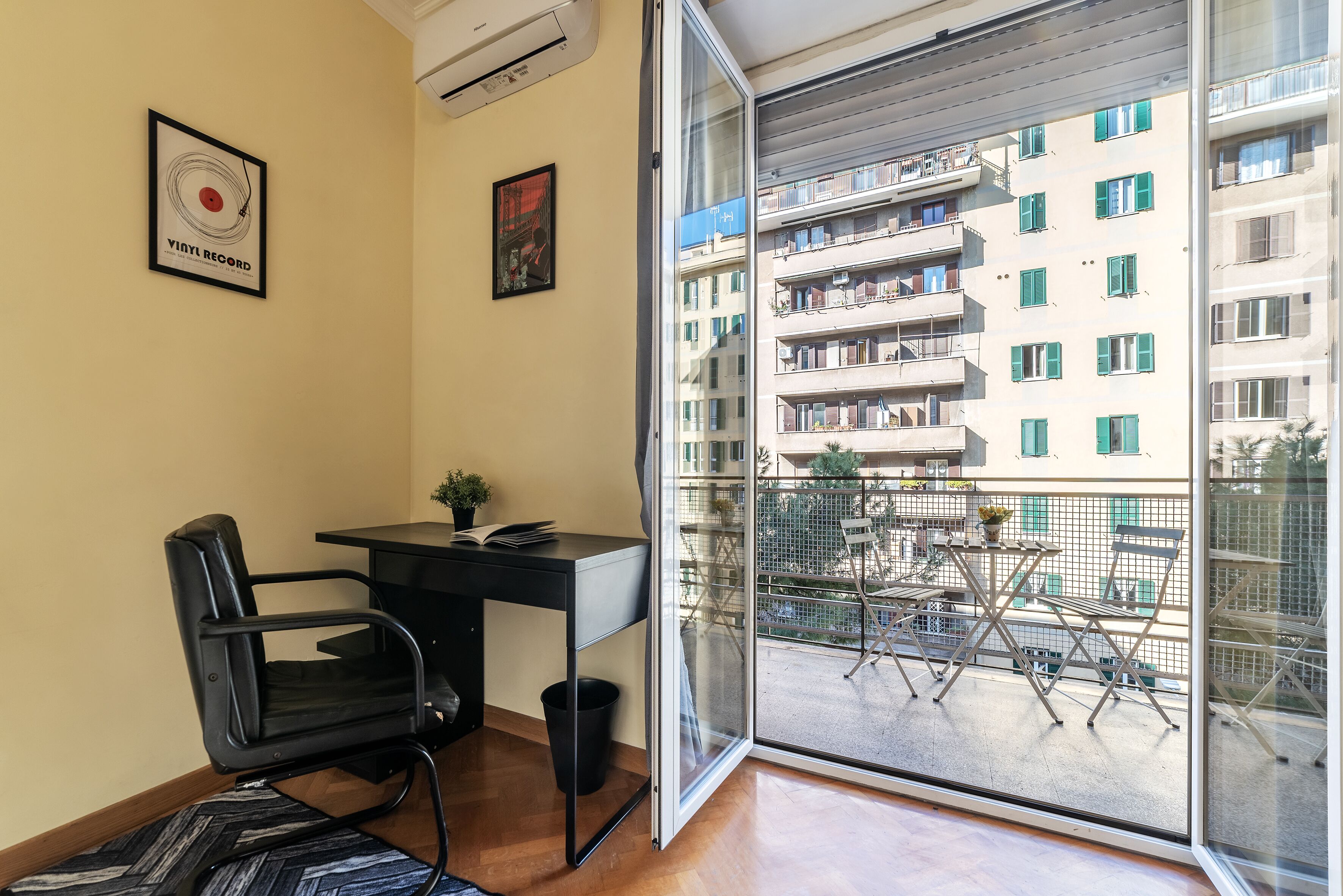 A room with a desk, chair, and a large window overlooking a balcony with outdoor furniture.