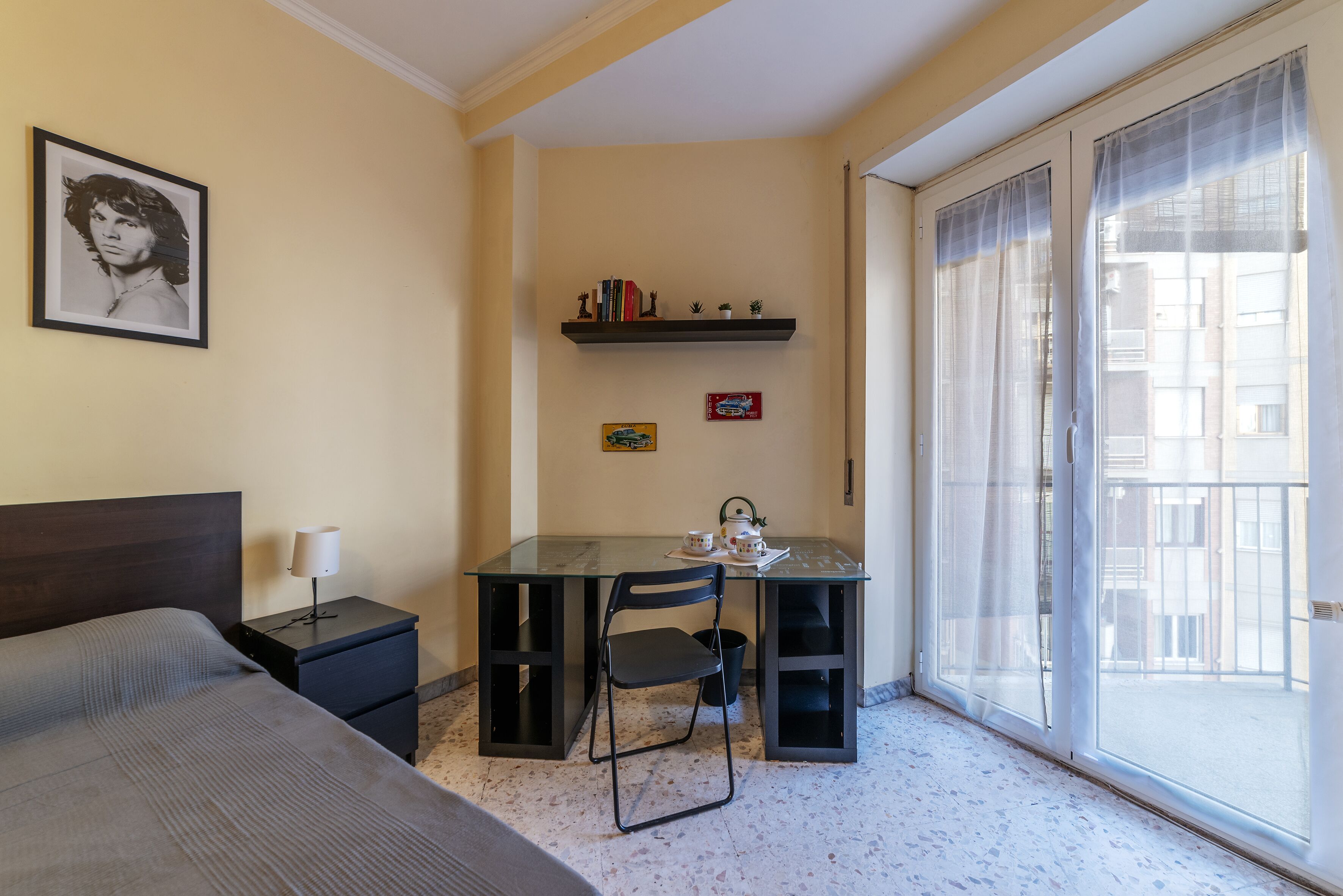 Une chambre à coucher avec un lit, un bureau avec une chaise, une petite table avec une théière et une fenêtre donnant sur des immeubles.