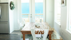 Un comedor con una mesa de madera y sillas de metal blanco, una araña y vista al océano a través de una puerta.