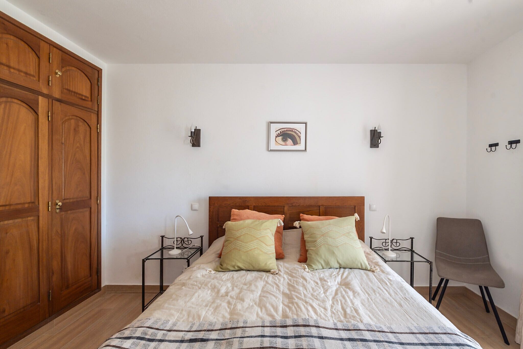 Un dormitorio con un armario de madera, una cama con dos almohadas, mesitas de noche con lámparas y una silla.