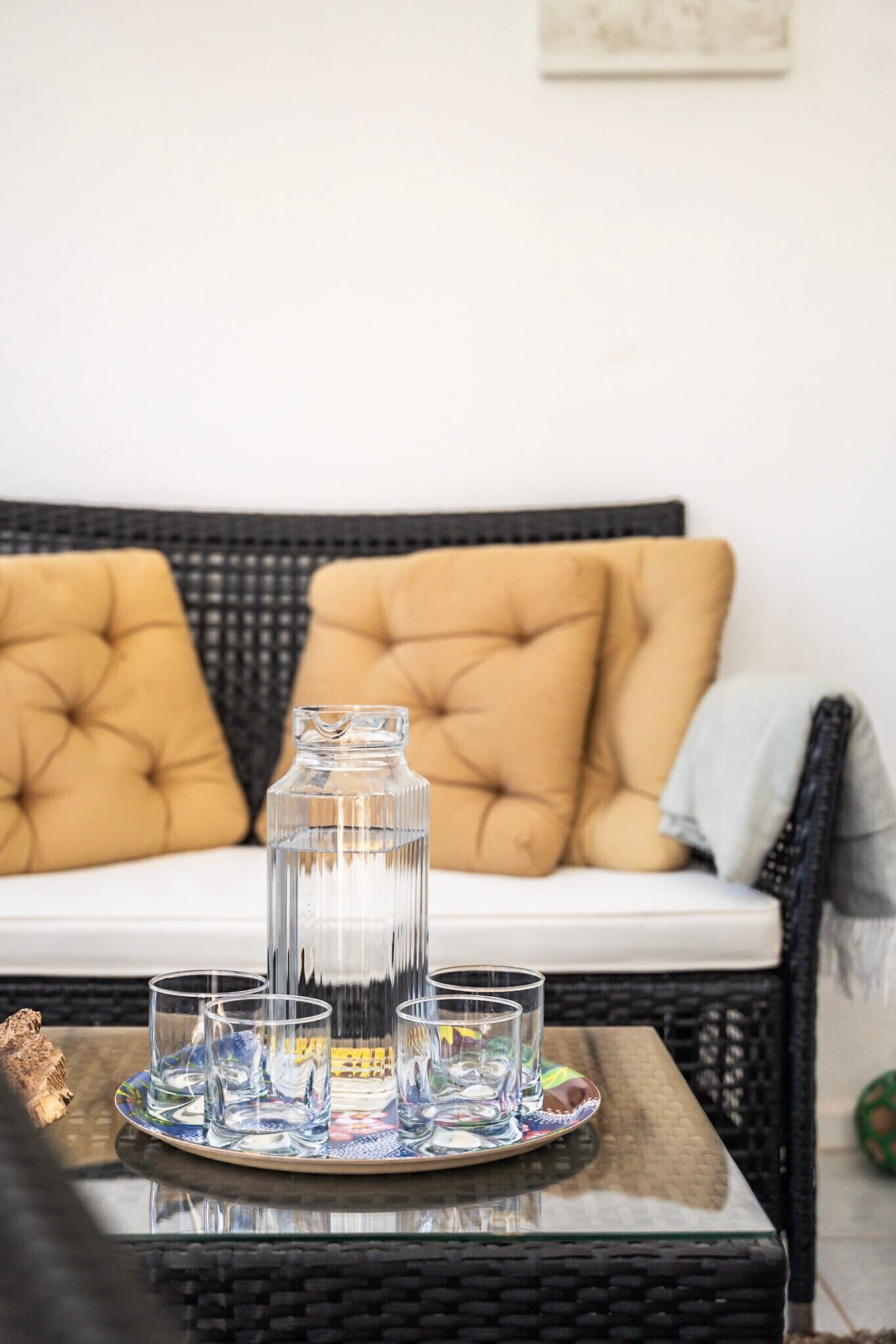 Une carafe en verre, posée sur un plateau, est entourée de quatre verres sur une table devant un canapé orné de coussins jaunes.