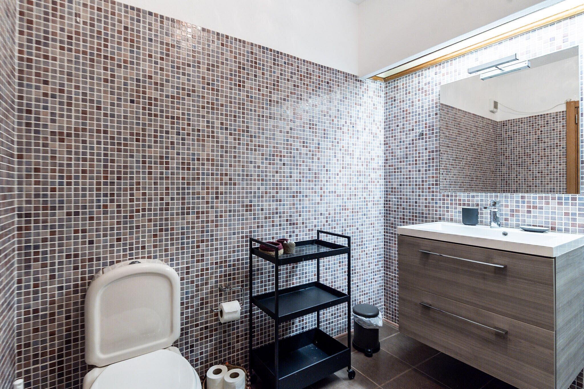 Une salle de bain avec des murs recouverts de mosaïques, des toilettes, une étagère noire où sont rangés les articles de toilette et un meuble-lavabo avec un miroir.