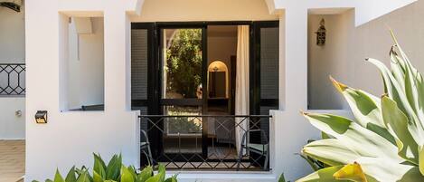 Un edificio blanco con un balcón, una puerta con contraventanas abiertas y un ventanal pequeño.