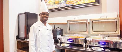 A buffet setup with a chef standing in front, a refrigerator, and two self-service beverage dispensers.
