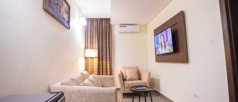 A hotel room with a sofa, a chair, a small table, a TV, an air conditioning unit, and a painting on the wall.