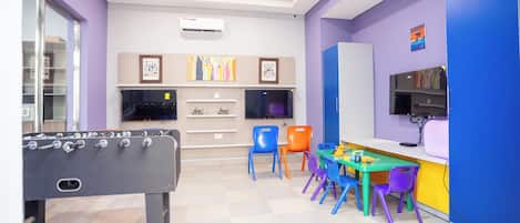A room with a foosball table, colorful chairs, a small table with items, and a TV on a stand.