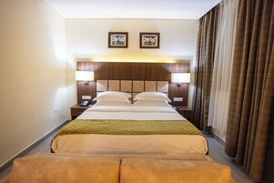 A hotel room with a large bed, two bedside lamps, a headboard with a light panel, a telephone, and a window with curtains.