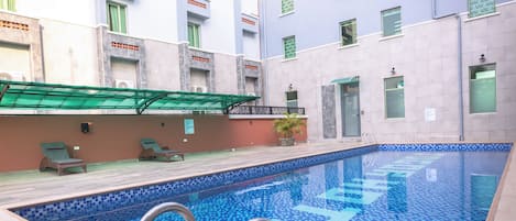 Une piscine avec un sol carrelé et un escalier en métal. On y trouve des chaises longues et un pot de fleurs sur la terrasse.